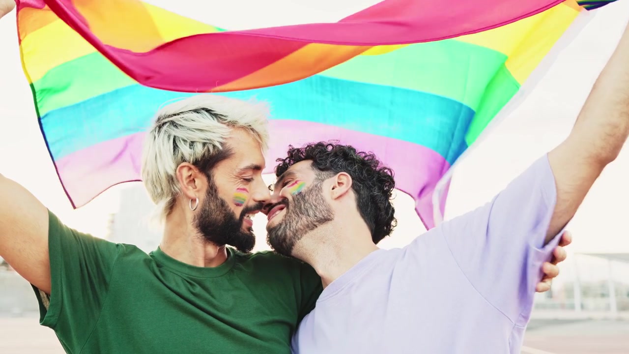はためくレインボーフラッグの下でキスをする幸せそうな同性カップル。
