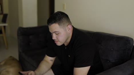 una toma estrecha de un joven latino de piel quejosa sentado en su sofá de la sala de estar usando su teléfono inteligente y sonriendo mientras conversa con la gente
