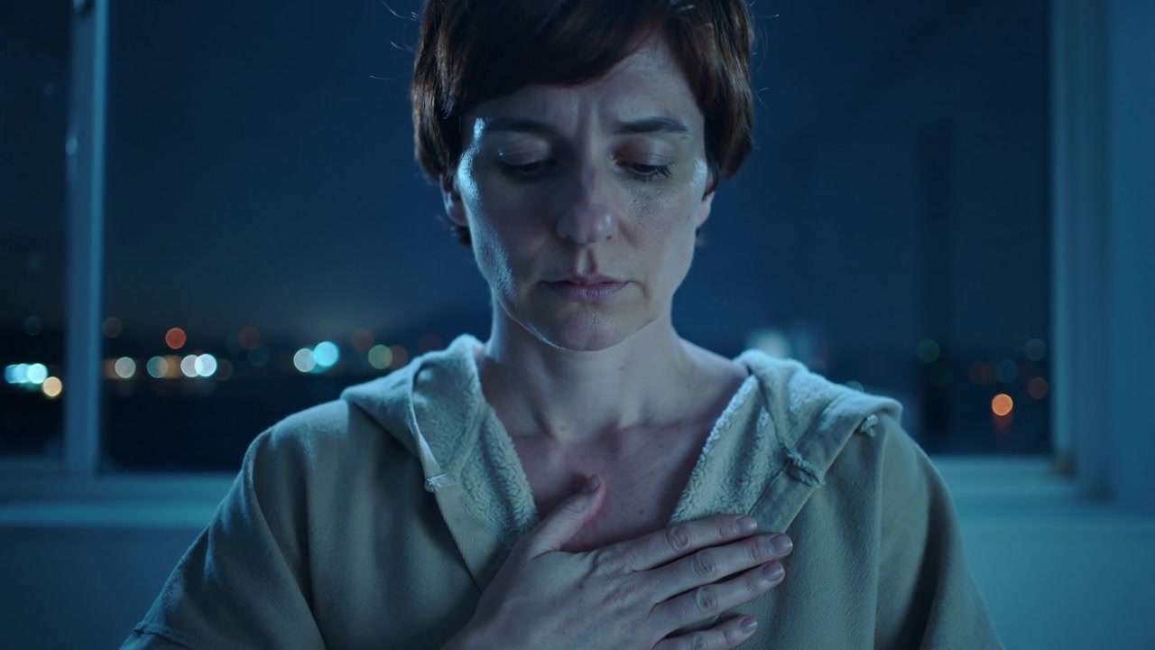 Donna che abbassa lo sguardo, alzando la mano destra e posizionando il palmo sullo sterno per calmarsi, indossando una felpa con cappuccio, con le luci della città sullo sfondo.