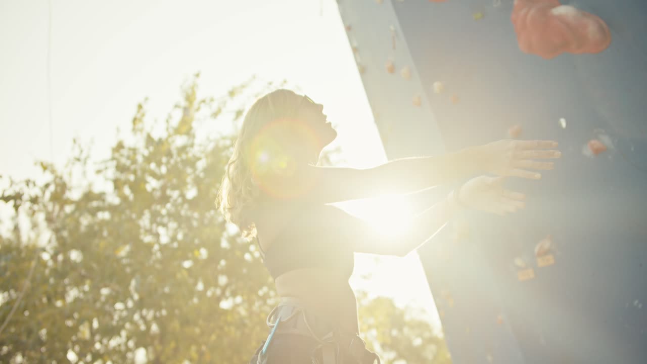 검은색 에 안경을 입은 금발 소녀가 여름의 은 날에 파란색 등반 벽을 오르기 전에 손과 어깨를 펴고 있습니다.