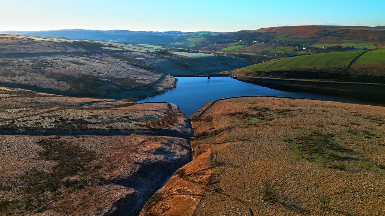 Luftoptagelser flyvende over gyldne hedeområder og rundt om en blå sø, vandreservoir i det fjerne