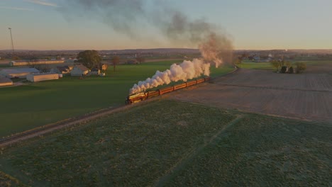 una vista aérea de las tierras de cultivo al amanecer, con graneros y granjas, con un tren de vapor acercándose soplando mucho humo en una mañana de otoño