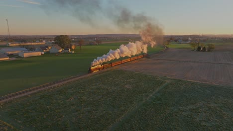 an aerial view of farmlands at sunrise, with barns and farms, with a steam train approaching blowing lots of smoke on an autumn morning