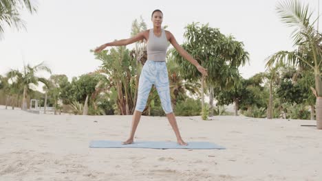 Happy-biracial-woman-practicing-yoga-standing-on-beach,-slow-motion