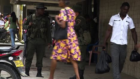 colombian military men standing on a busy sidewalk holding rifles while civilians walk past.