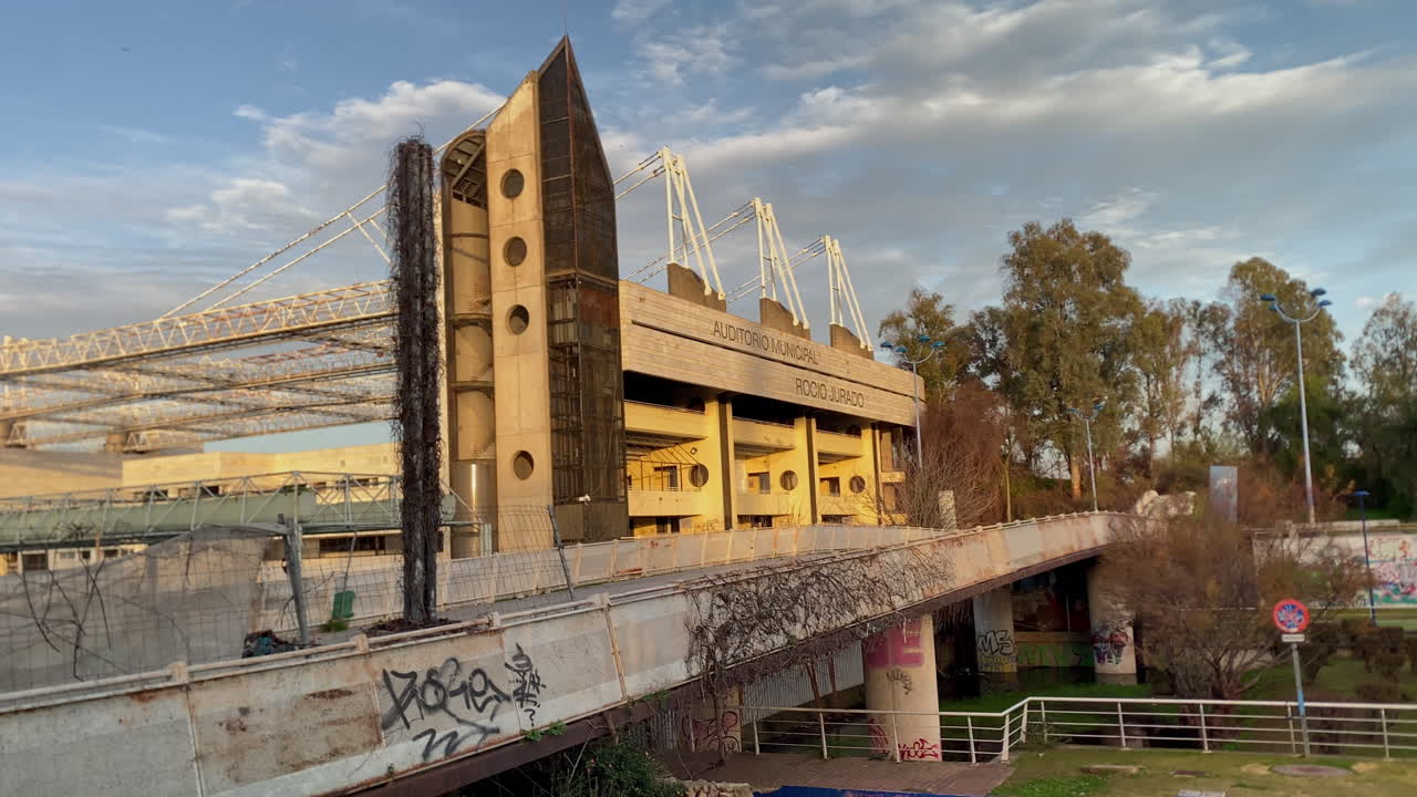 Vista urbana dell'Auditorium Rocio Jurado a Siviglia con una calda luce del tramonto, struttura moderna e passeggiata lungo il fiume circostante.