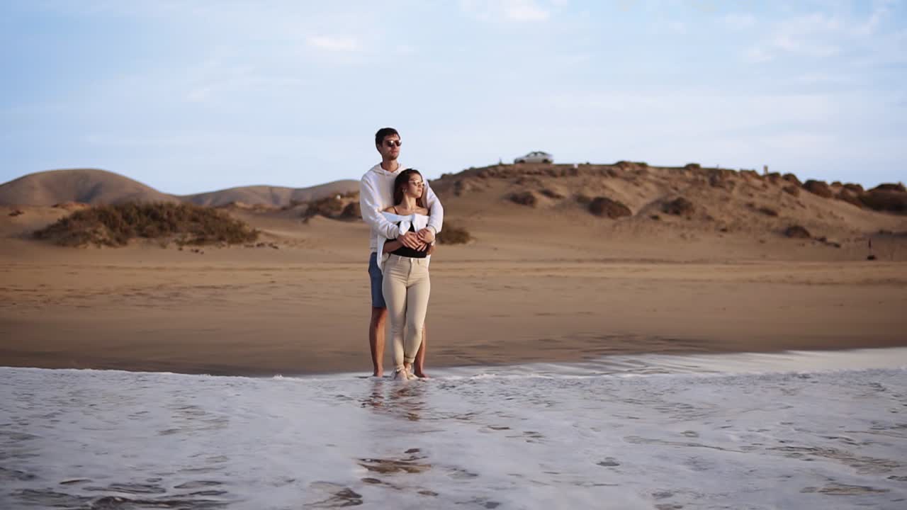 pareja de jóvenes que se unen de pie en la orilla de la playa de arena miran las olas del atardecer desde el océano. con gafas de sol y ropa casual. olas de espuma que vienen. vista frontal