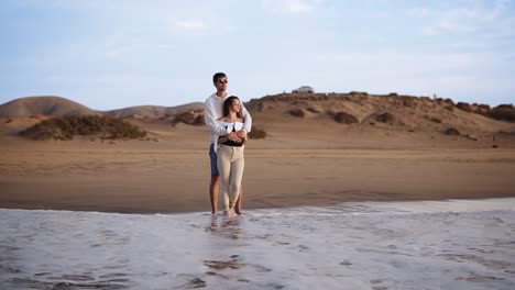Bonding-young-couple-standing-on-sandy-beach-shoreline-watch-sunset-waves-from-the-ocean.-Wearing-sunglsses-and-casual-clothes.-Foaming-wave-coming.-Front-view