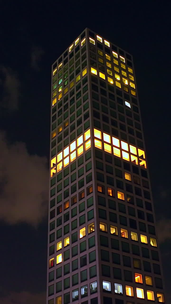 Top of the skyscraper with lights in the windows. Drone footage rising along the building at the backdrop of dark night sky with clouds. Vertical video