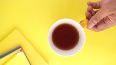 hand holding a mug of tea on a yellow background with notebooks and pencil.