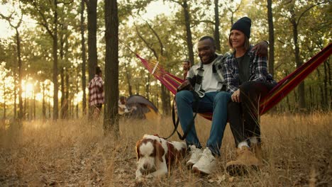 a happy couple, a man with black skin and a girl in a black hat in a plaid shirt, are sitting on a red hammock, next to them is their white-brown dog, and behind them is the rest of the group of people who are going on a hike and in a tent in a green sunny forest during the evening