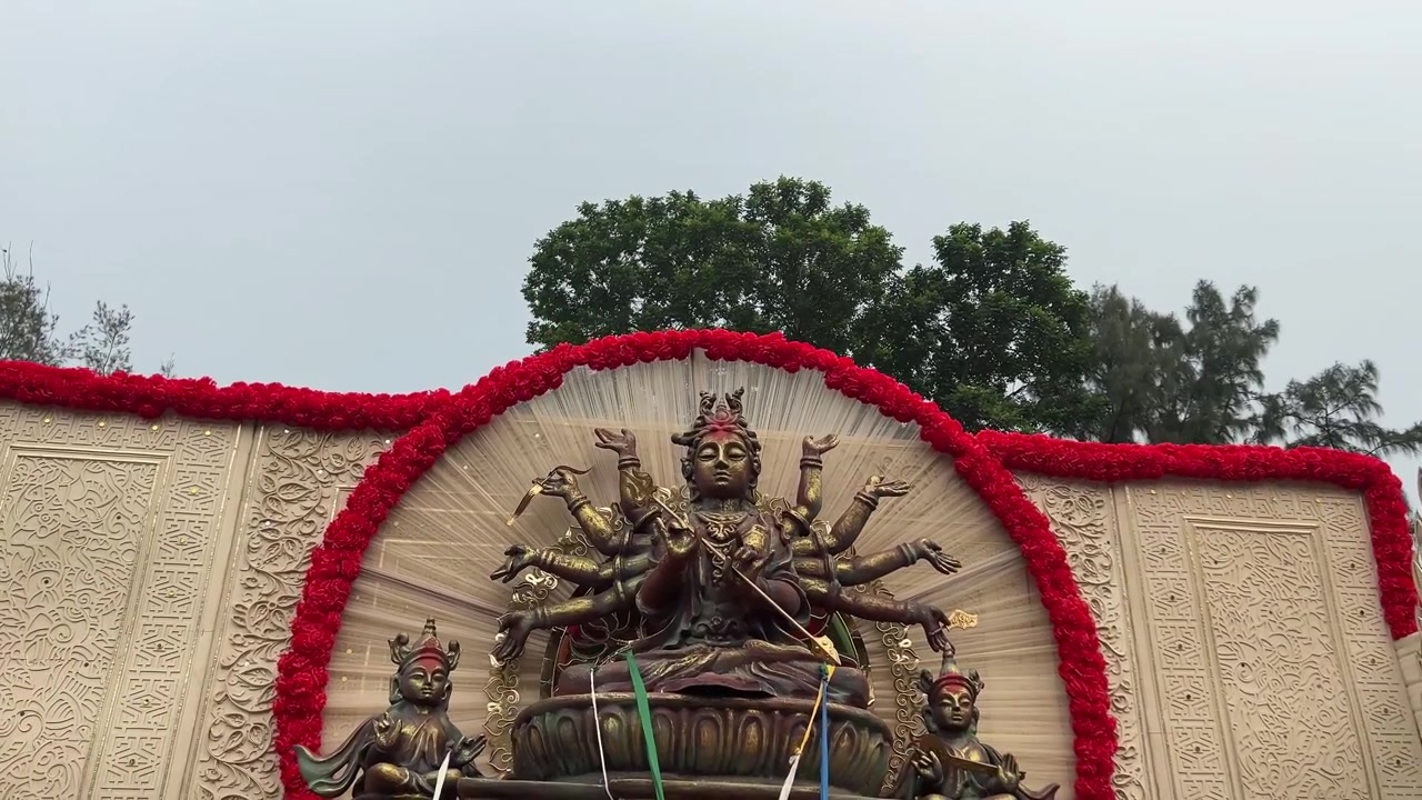 A dark golden idol of Maa Durga, with Laxmi and Saraswati, in a yellow ochre open pandal with a red flower border, sky, and trees in the background.