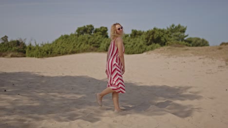 a beautiful young woman wearing a summer dress stands in sunny sand dunes surrounded by green forest