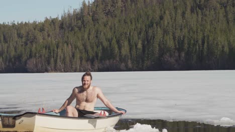 shirtless man sitting in a boat afloat on frozen lake in winter