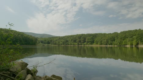 Woodland-landscape-with-calm-lake-and-reflections-of-cloudy-blue-skies