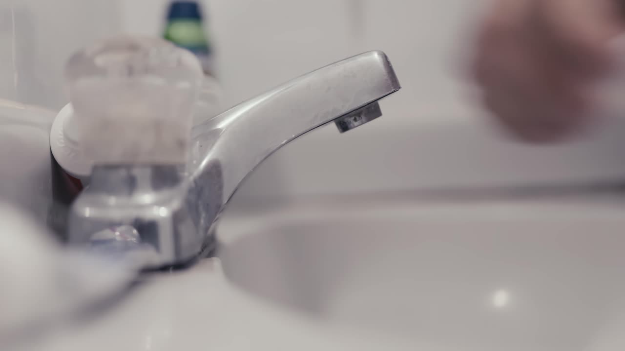 cerca de las manos de un joven latino blanco que se acerca a un fregadero del baño con su cepillo para ponerle un poco de pasta dental y procede a verter un poco de agua para humedecerlo