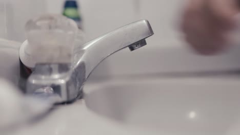 close-up-to-the-hands-of-a-white-Latino-young-man-approaching-to-a-bathroom-sink-with-his-brush-to-put-some-dental-paste-on-it-and-proceeds-to-pour-a-bit-of-water-to-wet-it