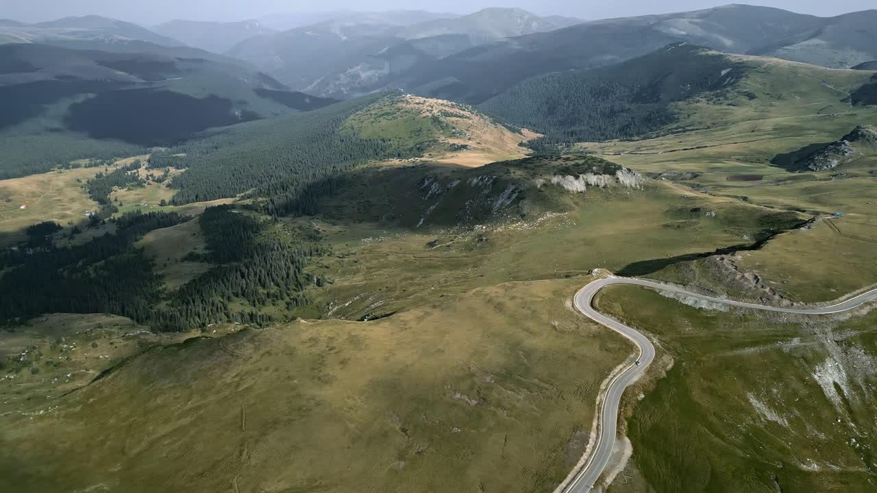 A breathtaking bird's-eye view of Transalpina in Romania, showcasing the contrasting shades of dense forests, golden hills, and a winding road snaking through the undulating landscape