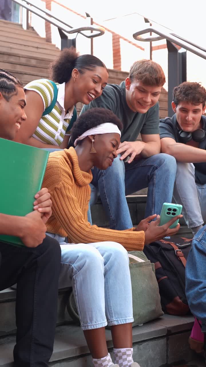 Gruppo eterogeneo di studenti che ridono mentre usano lo smartphone sulle scale del campus.