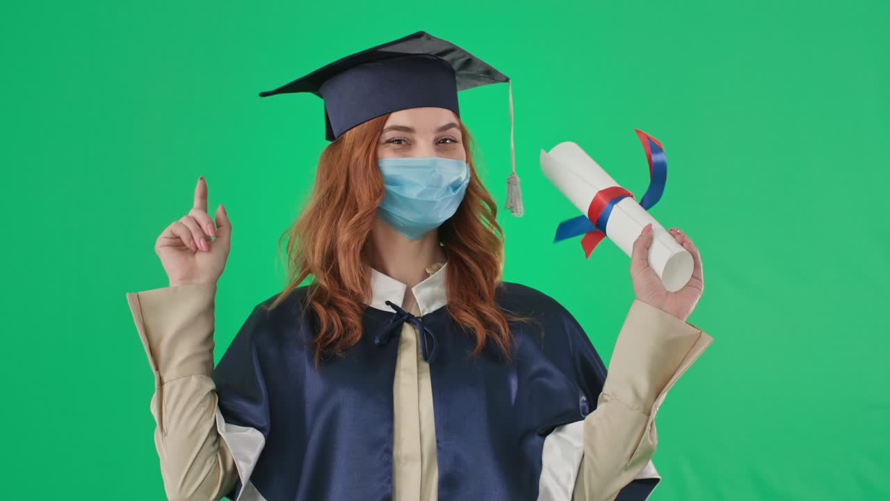 diploma online, laureato gioioso in maschera medica si rallegra per il certificato di istruzione ricevuto e indica le dita durante l'apprendimento a distanza sfondo schermo verde, chiave croma