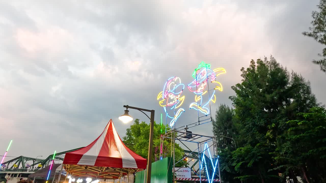 Colorful neon lights illuminate a bustling market scene under a cloudy sky in Bangkok, creating a lively atmosphere