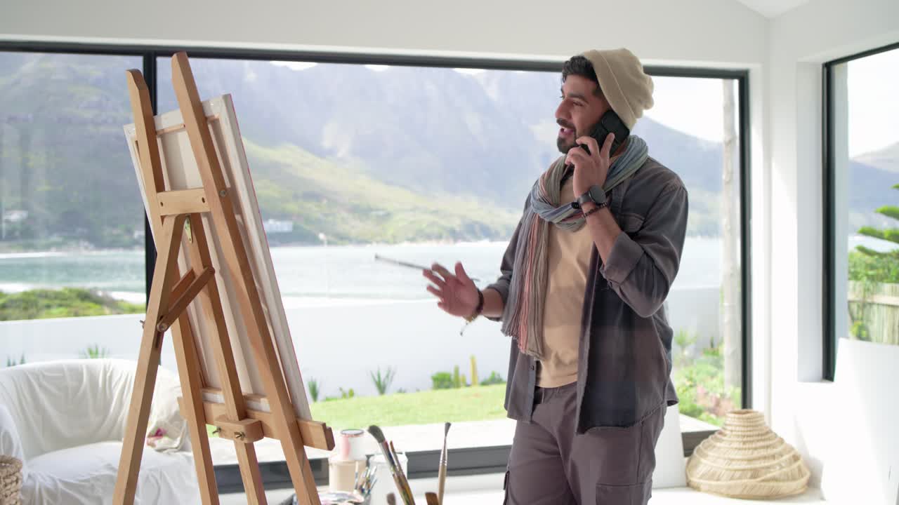 En indisk mann besvarer en telefonsamtale mens han ferdigstiller et maleri ved staffeli, med lue, i et studio ved en innsjø.