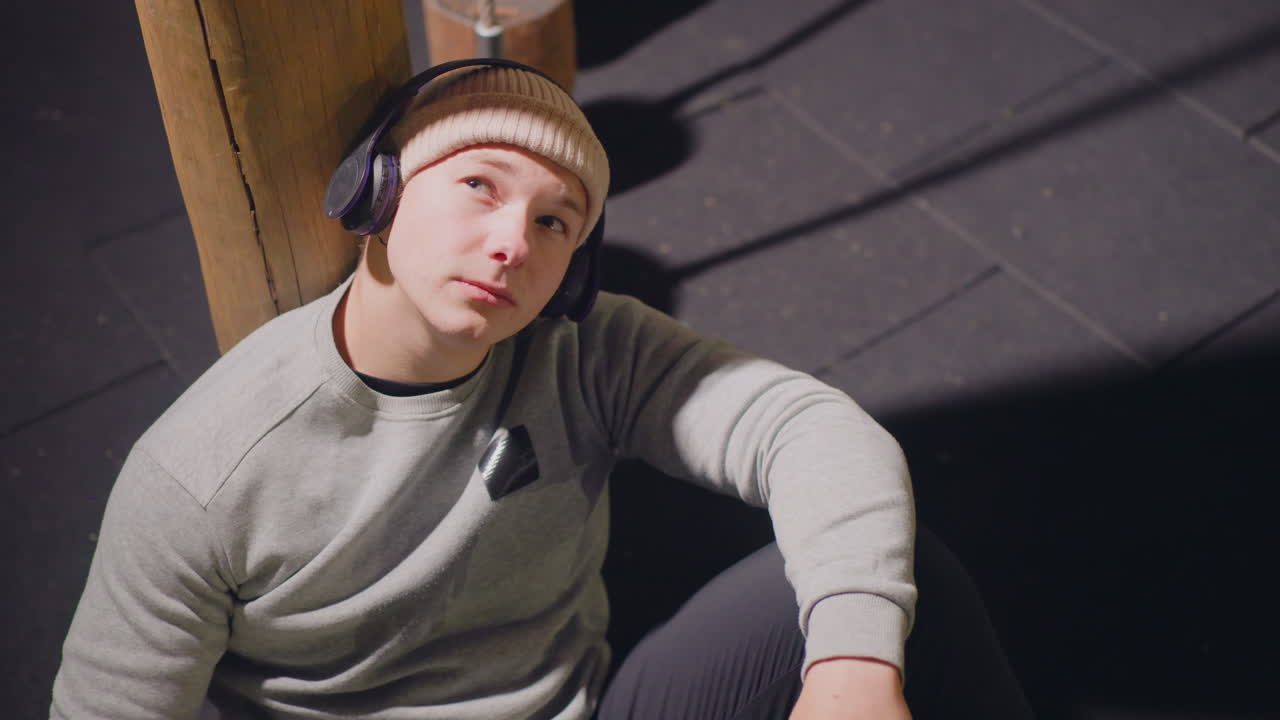 Jovem com fones de ouvido sentado perto de um pilar de madeira no chão iluminado de uma academia, usando gorro de tricô e suéter, com uma expressão relaxada, mas pensativa. Sombras suaves e destaques quentes criam um clima intimista.