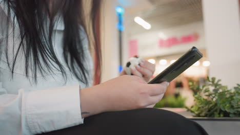 young woman with headset seated indoors operating digital device while gently squeezing stress ball, wearing glasses with long dark hair, blurred storefront in background