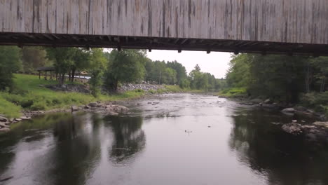 Aerial-shot-flying-above-the-Piscataquls-River-and-underneath-the-covered-wooden-Lowes-Bridge-in-Maine