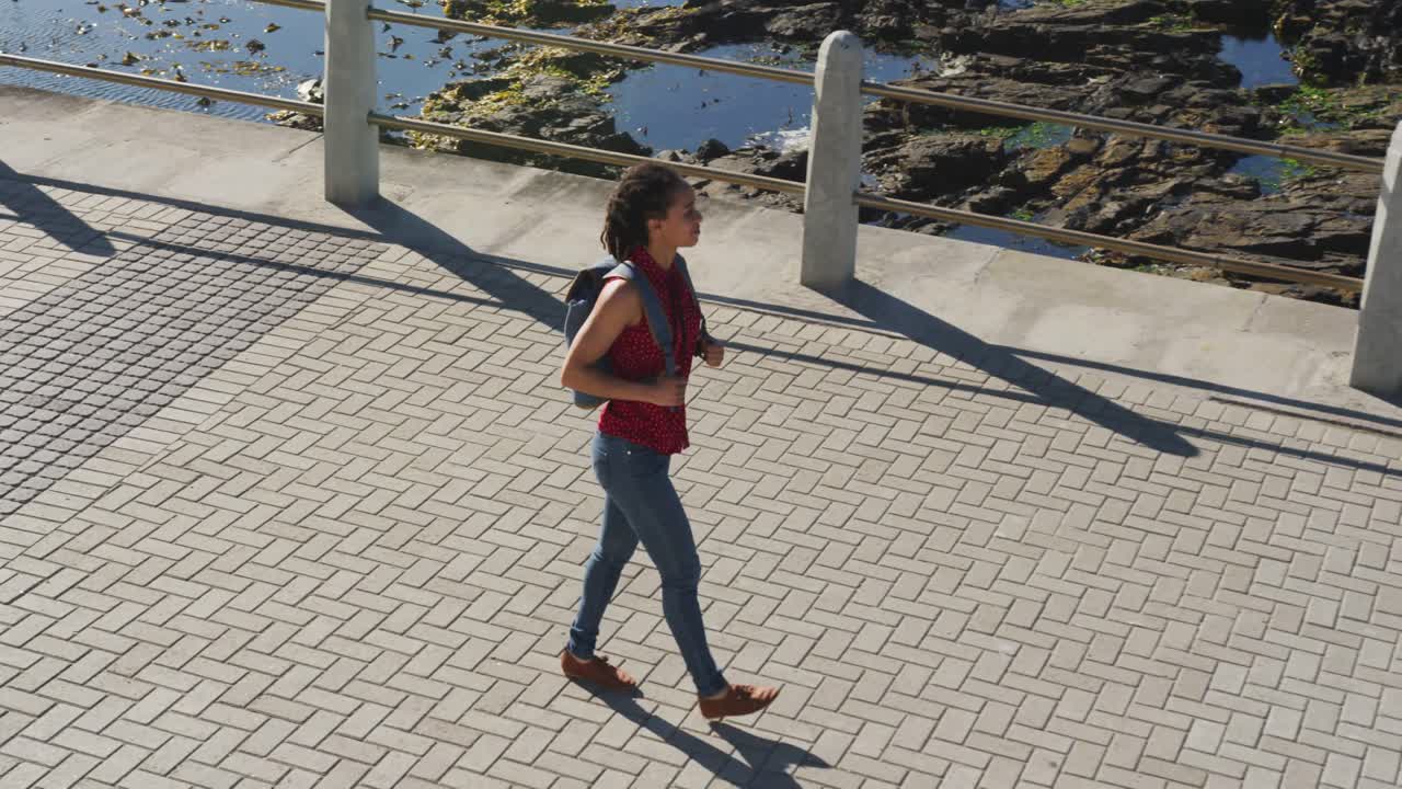 African american woman wearing backpack and walking on promenade by the sea