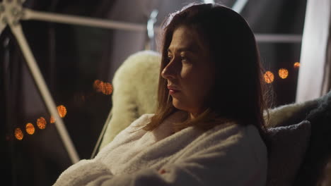 upset woman sits in fluffy armchair thinking over bad news on glass balcony with wide panoramic windows. young lady with dark hair suffers from depression