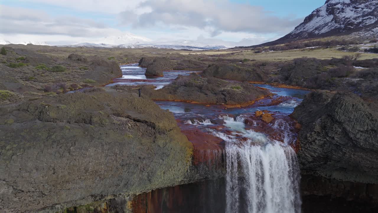 Person standing at the edge of Salto del Agrio waterfall with colorful volcanic rocks and snowy mountains in background