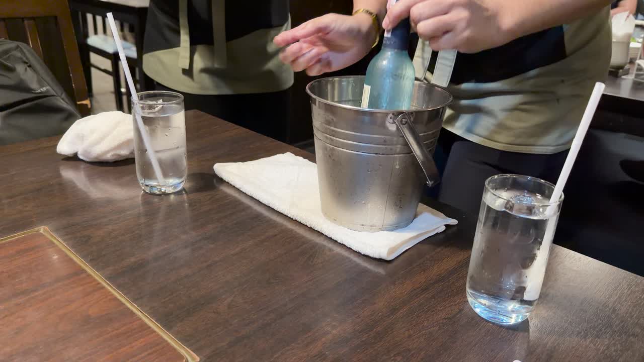 Server sets glass and chilled sake bottle in ice bucket on wooden table, natural light