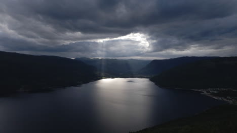 Aerial-shot-of-lake-in-between-the-mountains-with-sunrays-coming-out-from-the-clouds