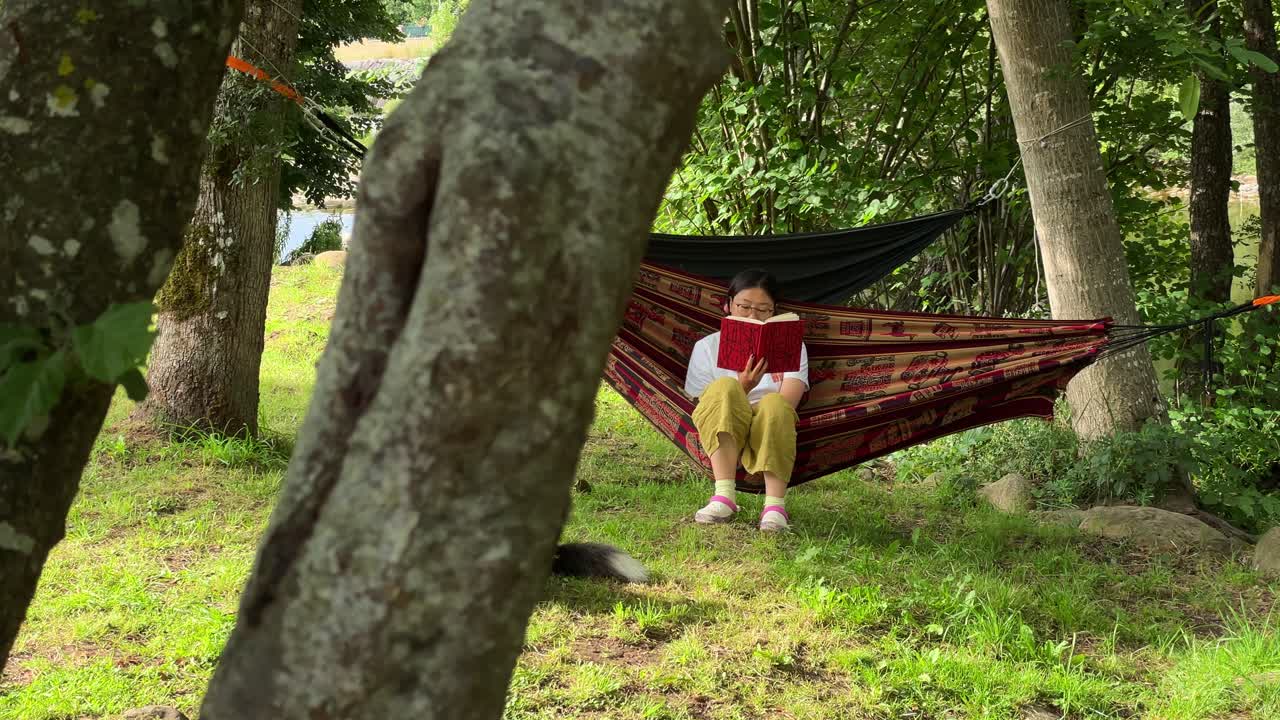 una chica asiática leyendo un libro en una hamaca junto al río, con su perro
