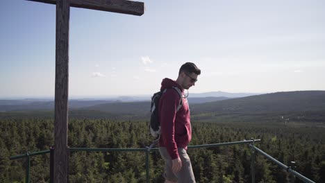 young tourist man standing at the top of hill and looking around, wooden cross is next to him