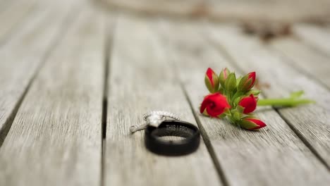 A-round-brilliant-cut-diamond-ring-set-up-on-top-of-a-black-wedding-band-on-a-light-wooden-table-with-a-red-budding-flower-is-pushed-into-focus