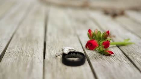 a round brilliant cut diamond ring set up on top of a black wedding band on a light wooden table with a red budding flower is pushed into focus