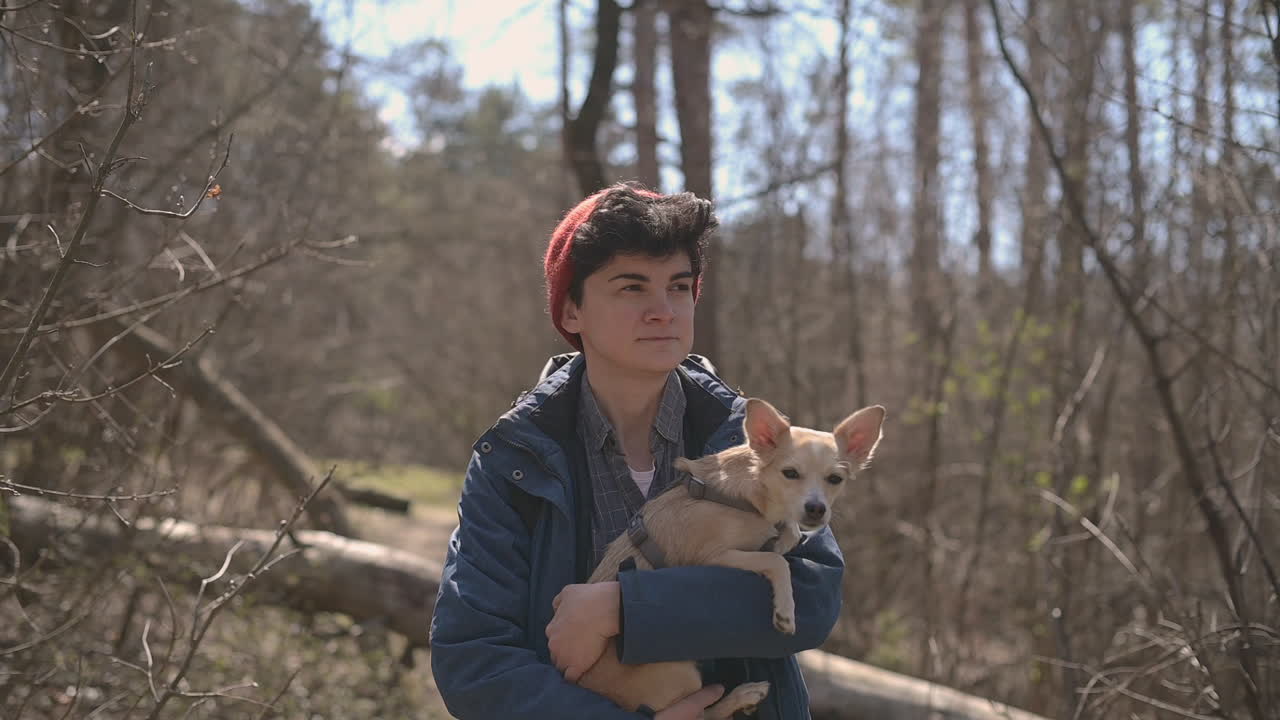 짧은 머리를 가진 젊은 여성이 작은 개를 안고 숲을 산책한다 1