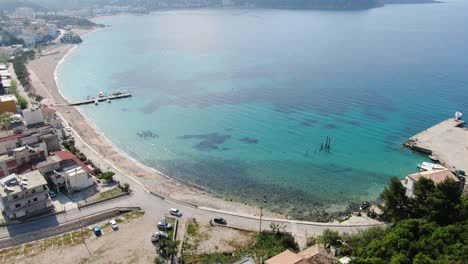 Drone-view-in-Albania-flying-over-a-beach-with-crystal-clear-blue-water-ocean,-buildings-on-the-harbor-and-green-island-on-a-sunny-day