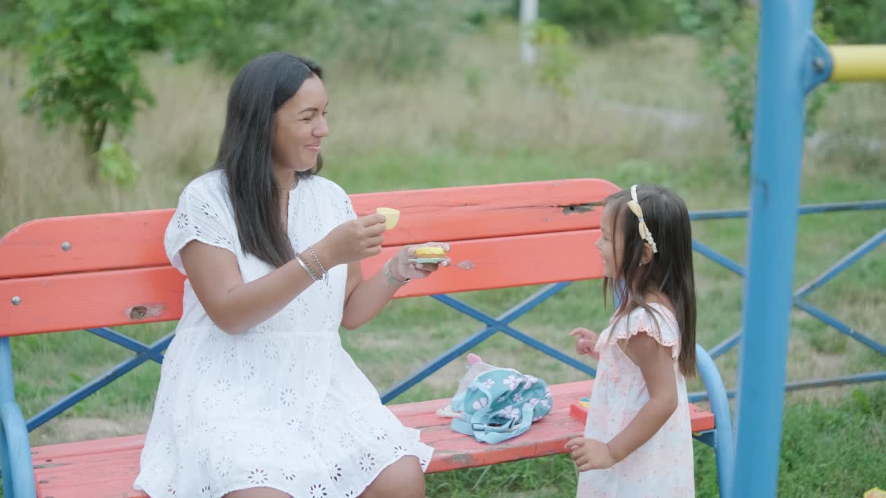 임신 한 어머니 와 어린 딸 은 나무 와 초록색 으로 둘러싸인 공원 의 놀이터 에서 함께 놀이 를 즐긴다