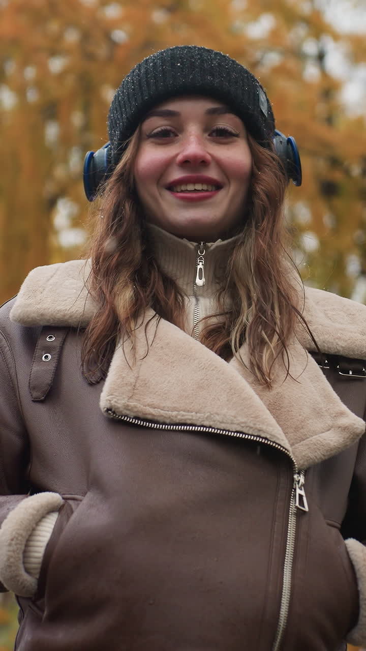 Smiling female wearing black knit cap and brown shearling jacket with hands in her pockets outdoors during cool autumn day, enjoying the moment in nature