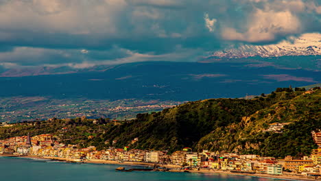 Aerial-drone-shot-soaring-above-Taormina,-Sicily,-Italy-with-enchanting-mountains-along-the-azure-sea-with-sunlight-shining-through-white-clouds