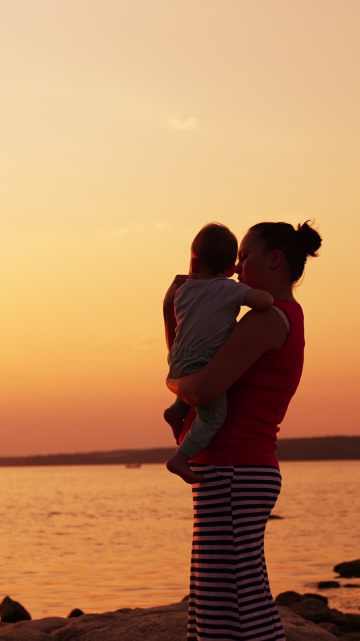 Caring and loving mother with her little baby in hands standing near the river. Family enjoying time at the nature at sunset. Vertical video