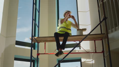 A-worker-eats-a-sandwich-on-the-construction-site,-sits-high-on-scaffolding