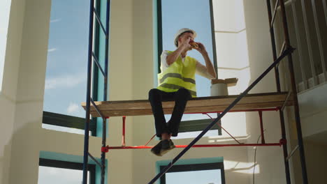 a worker eats a sandwich on the construction site, sits high on scaffolding