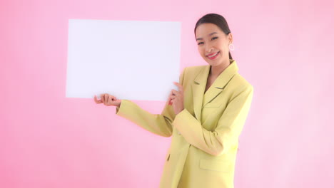 mujer de negocios asiática sosteniendo una hoja en blanco del cartel de papel con una expresión de cara feliz en el fondo del estudio rosa