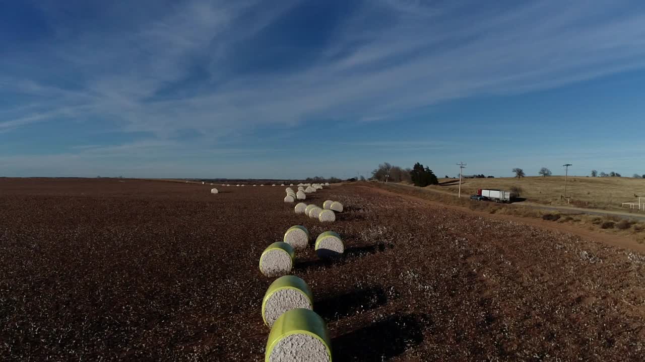 toma aérea de un dron en movimiento de una granja de algodón del medio oeste con pacas frescas de algodón cosechado envuelto en material amarillo brillante contra un semi camión azul de cielo abierto en la carretera