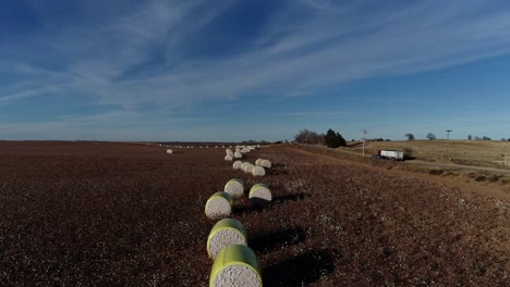 toma aérea de un dron en movimiento de una granja de algodón del medio oeste con pacas frescas de algodón cosechado envuelto en material amarillo brillante contra un semi camión azul de cielo abierto en la carretera