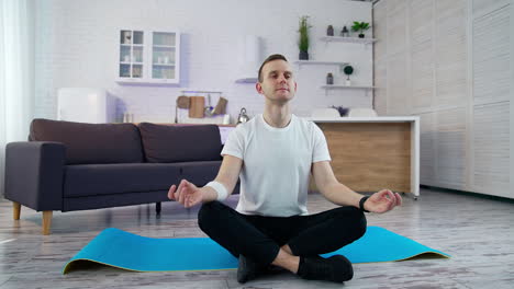 young man sitting in yoga pose in the kitchen. guy in white t-shirt doing relax exercises after workout while sitting on a mat at home.